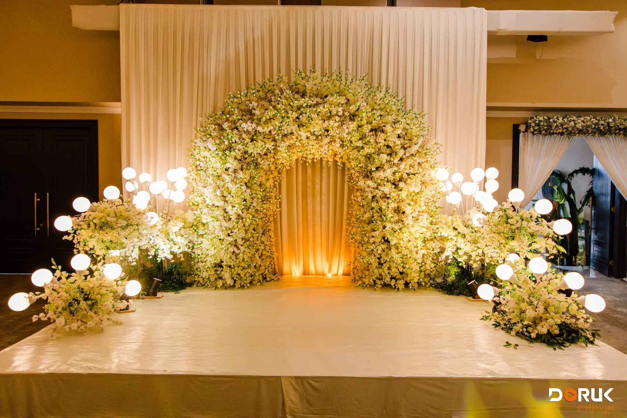 Bühne im Saal mit weißem Blumenbogen, leuchtenden Lichtkugeln und golden beleuchtetem Vorhang